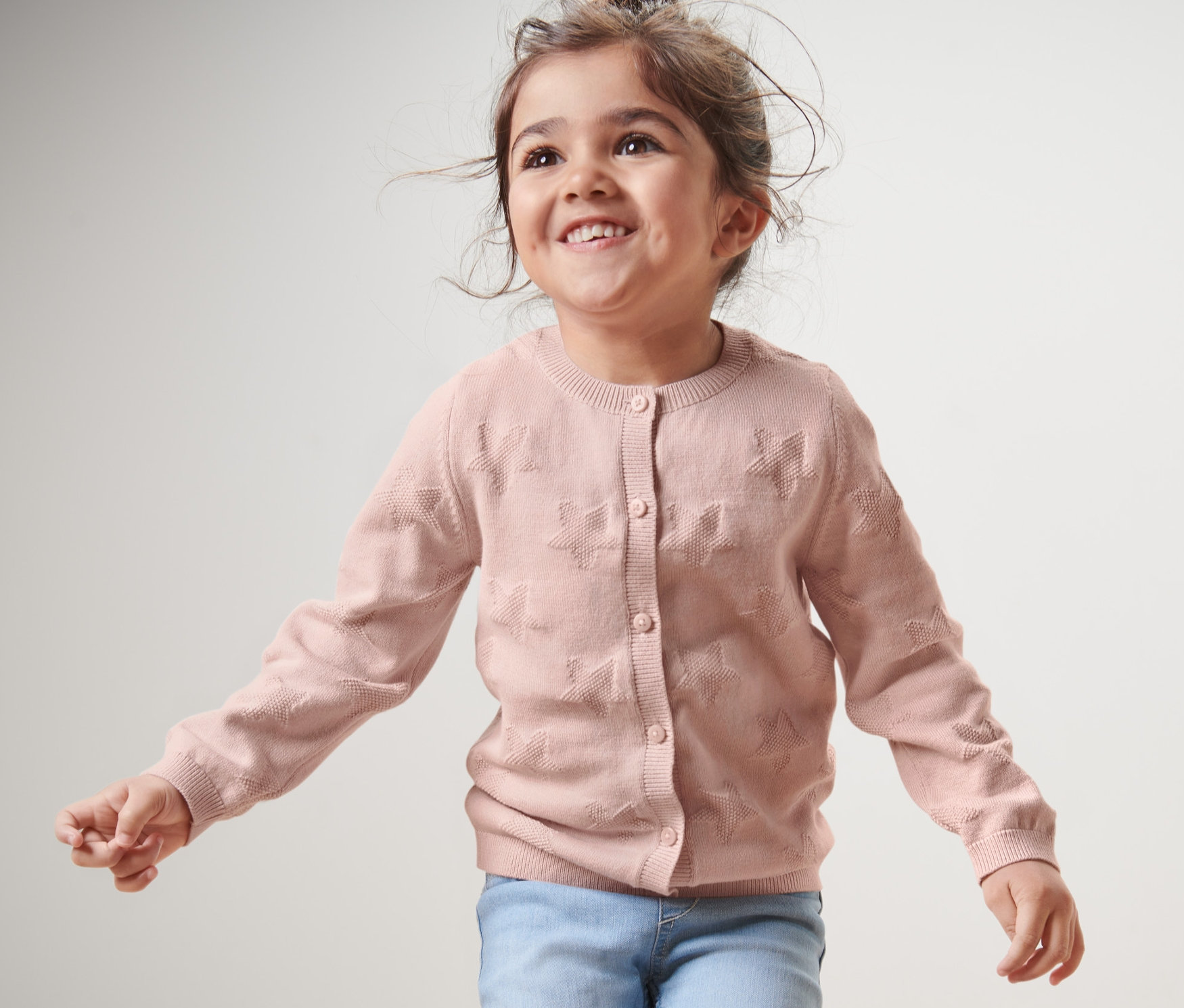Une fille souriante porte une jaquette en maille pour enfant et un jean.