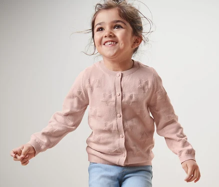 Une fille souriante porte une jaquette en maille pour enfant et un jean.