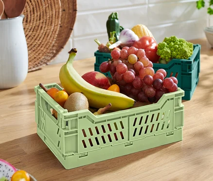 Une boîte de rangement pliable verte avec une banane, une pomme, un kiwi et des raisins. À l'arrière-plan, une boîte de rangement pliable verte avec des légumes.