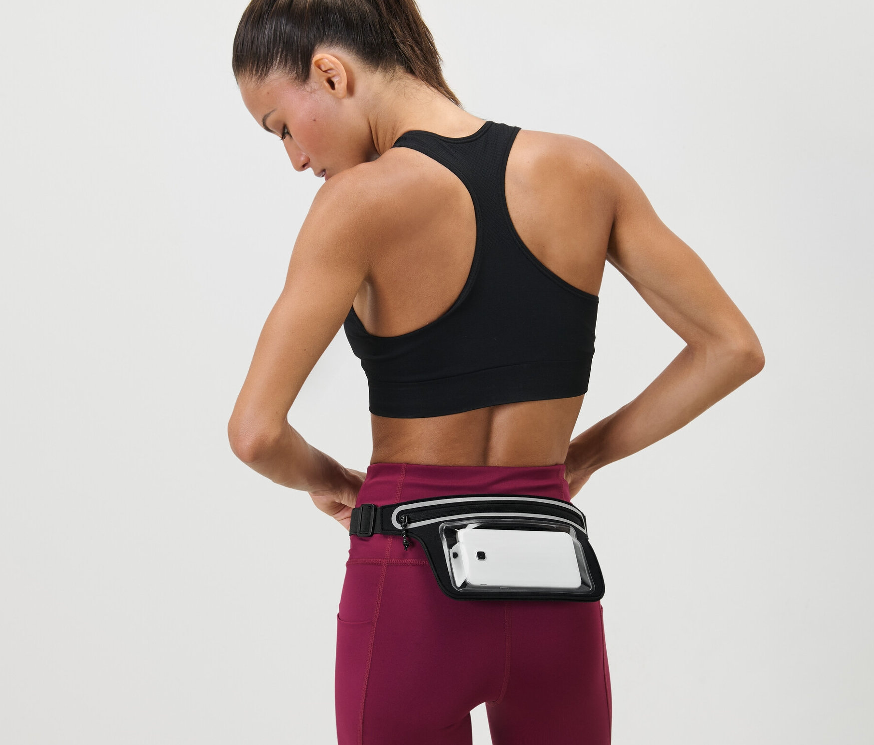 Femme ajustant une Ceinture de course pour smartphone autour de sa taille. Elle porte un soutien-gorge de sport et des leggings.
