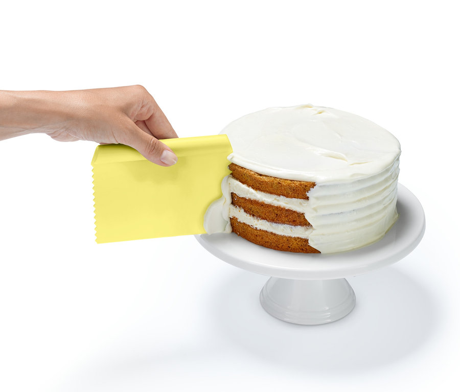Une main utilise un grattoir jaune du lot d'ustensiles de pâtisserie pour appliquer du glaçage blanc sur un gâteau à trois étages sur un socle blanc.