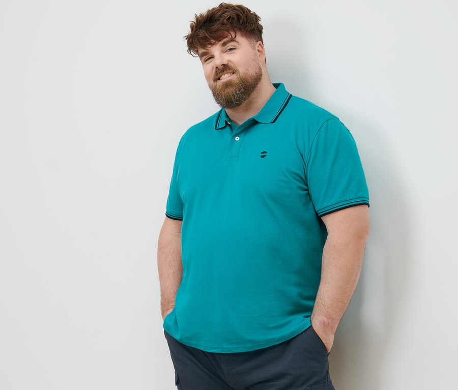 Un homme aux cheveux bruns se tient debout, les mains dans les poches de son short cargo bleu marine. Il porte un polo en piqué bleu turquoise.