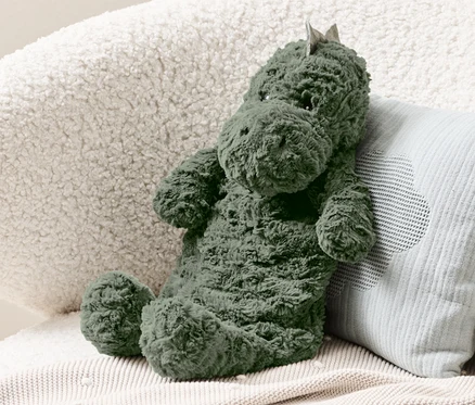 Une bouillotte dinosaure verte est posée sur un fauteuil crème à côté d'un coussin bleu clair.