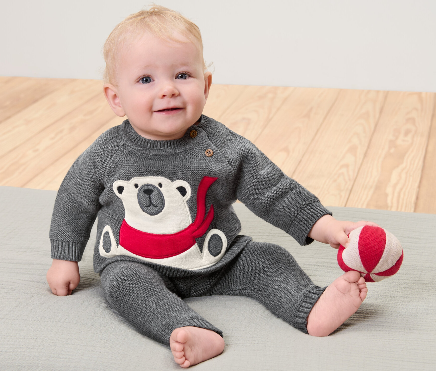 Un enfant est assis sur un tapis gris et joue avec une balle rouge et blanche. Il porte un pull en maille enfant gris avec un motif d'ours polaire.