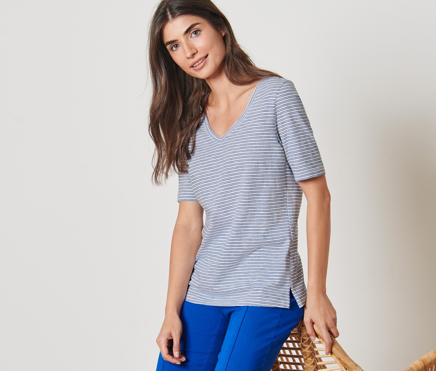 Une femme appuyée sur un fauteuil en rotin porte un t-shirt rayé et un pantalon bleu.