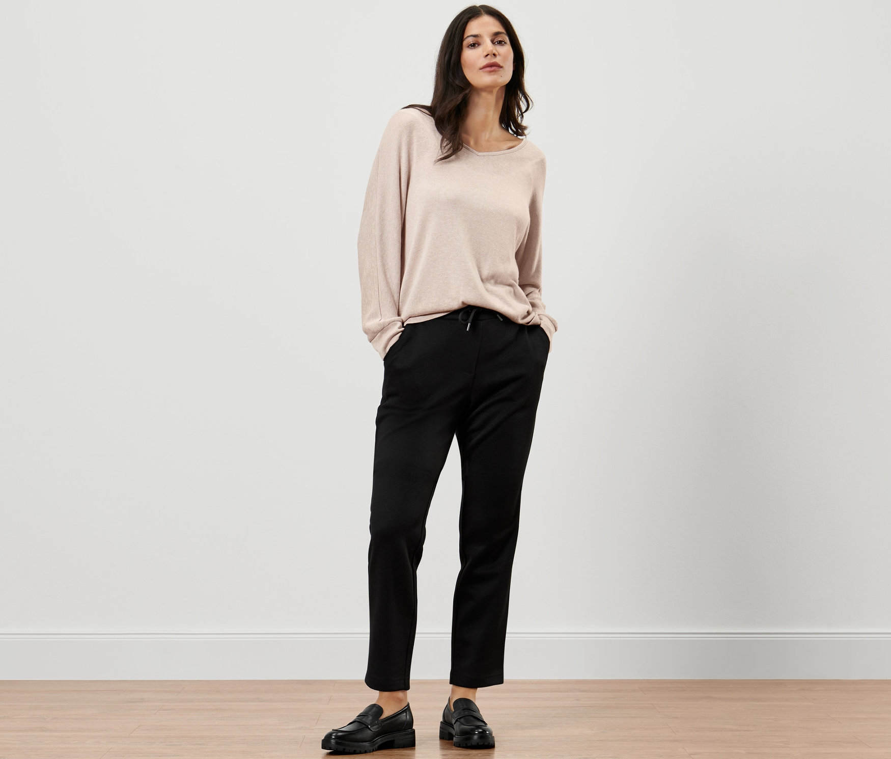 Une femme pose avec un pull beige et un pantalon de jogging noir.