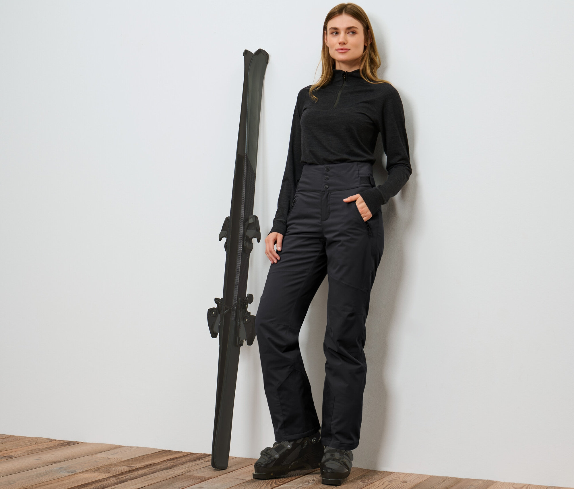 Femme posant à côté de skis en pantalon de ski taille haute noir.