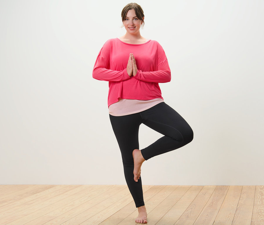 Femme portant un t-shirt de sport 2 en 1 rose et un legging de sport noir faisant la pose de l'arbre.