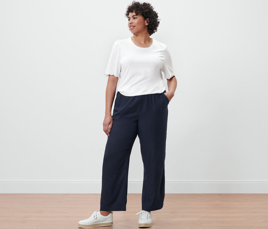 Une femme pose en pantalon tissé bleu foncé et chemise blanche à fronces.