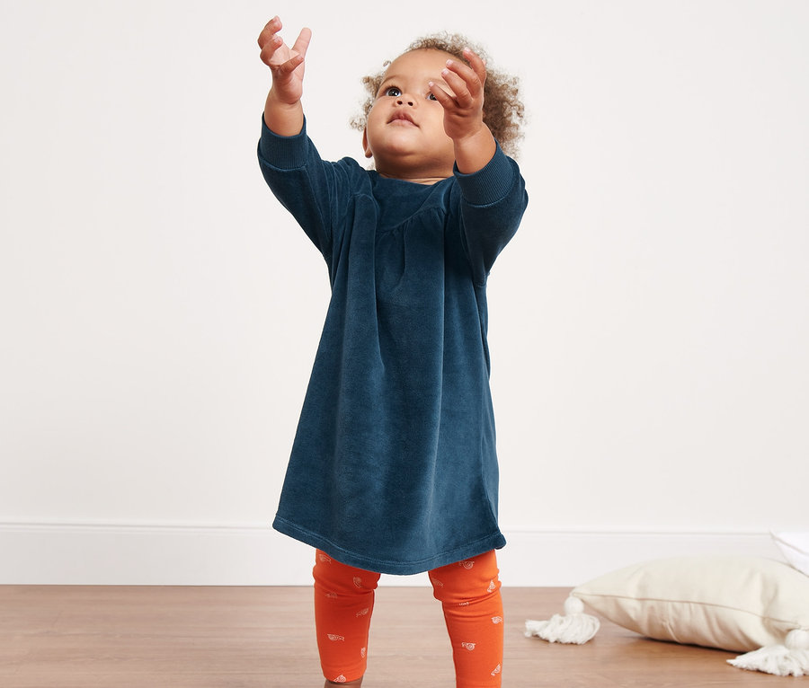 Une petite fille tend la main vers des bulles de savon. Elle porte une robe nicki pour bébé et des leggings orange.