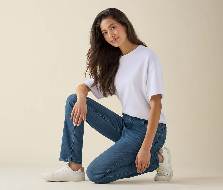 Une femme agenouillée sur un genou et souriante, elle porte un t-shirt blanc, un jean bleu et des baskets blanches.