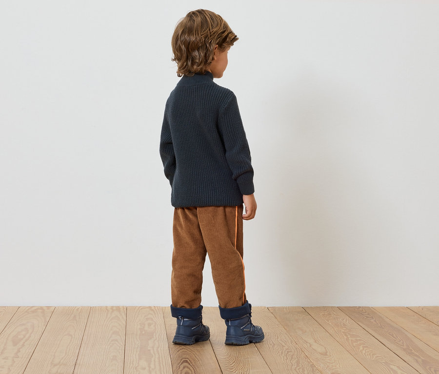 Un garçon se tient dos à la caméra, portant un pull bleu marine, un pantalon marron et des bottes bleues.
