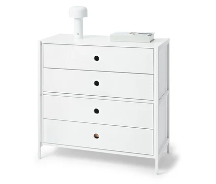 Commode blanche avec quatre tiroirs, sur le dessus une lampe et un livre.