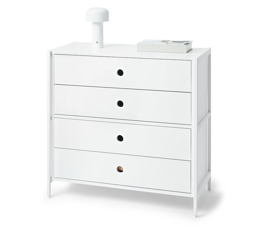 Commode blanche avec quatre tiroirs, sur le dessus une lampe et un livre.