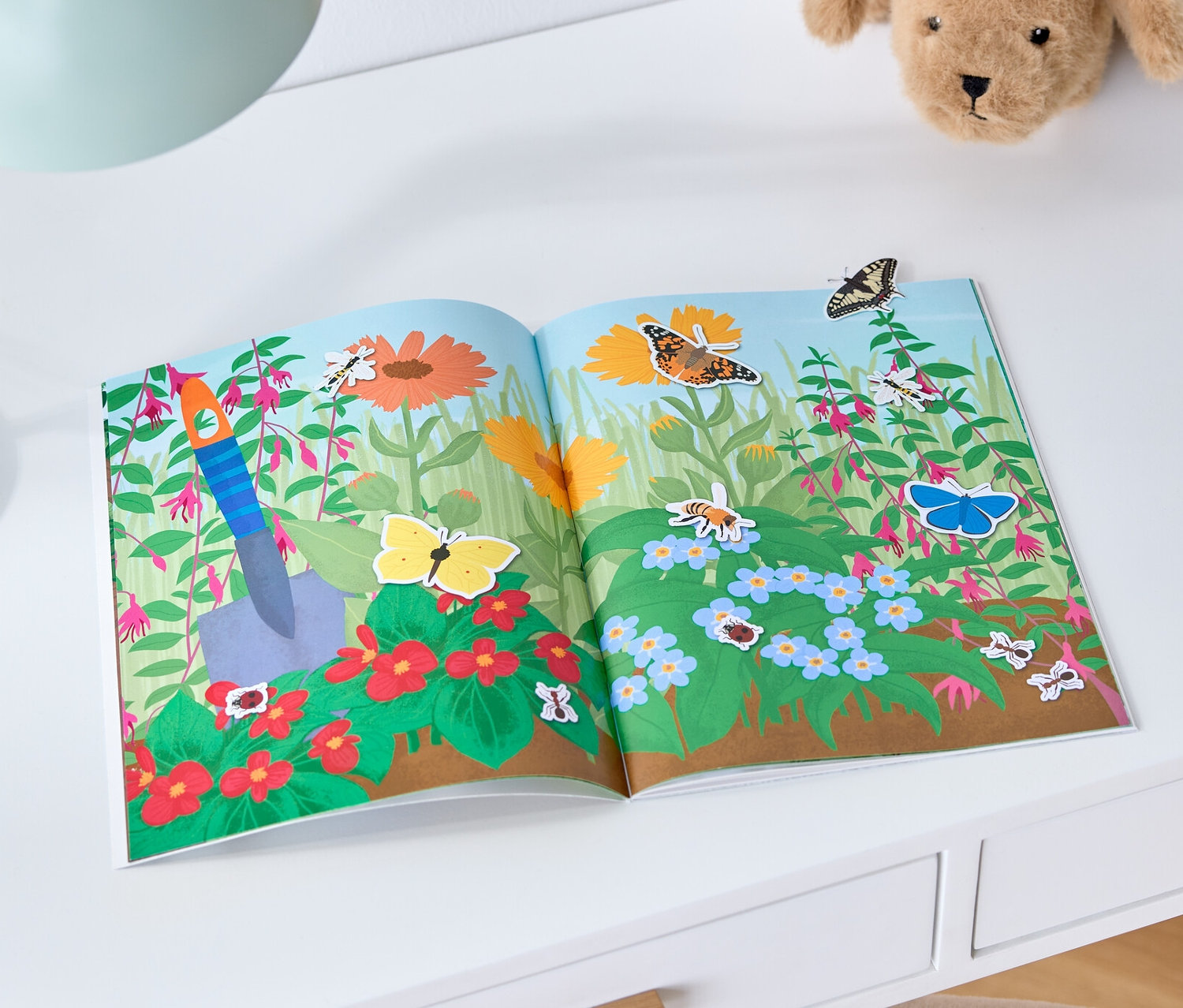 Sur une table blanche sont posés un livre d'autocollants ouvert « Les 4 saisons » (en allemand), une lampe et un chien en peluche.