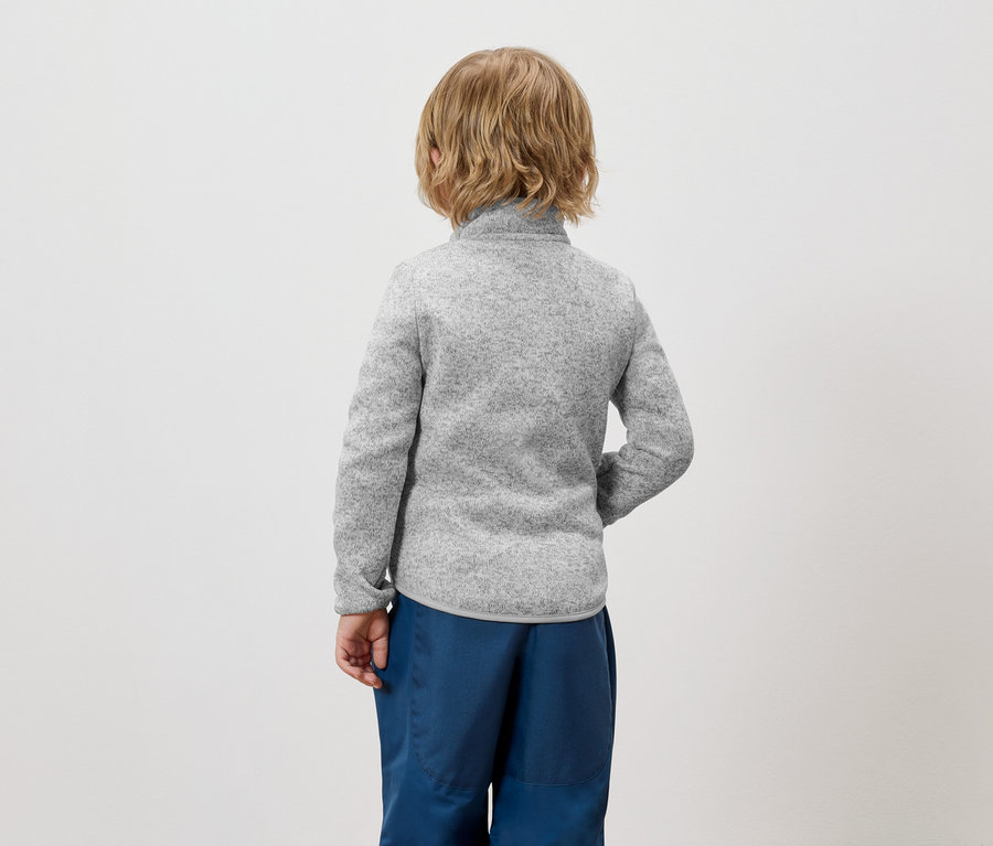 Un garçon aux cheveux blonds, vu de dos, portant un pull gris et un pantalon bleu.