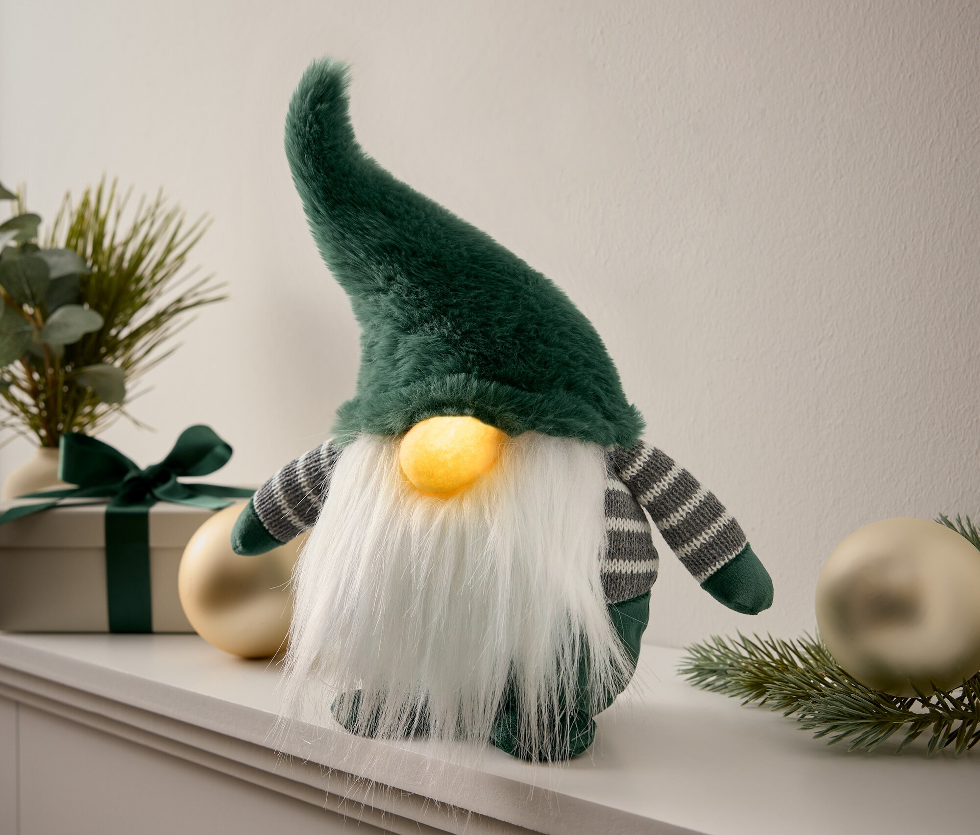 Un gnome en peluche vert avec une barbe blanche se tient sur une étagère à côté de décorations et d'un cadeau.