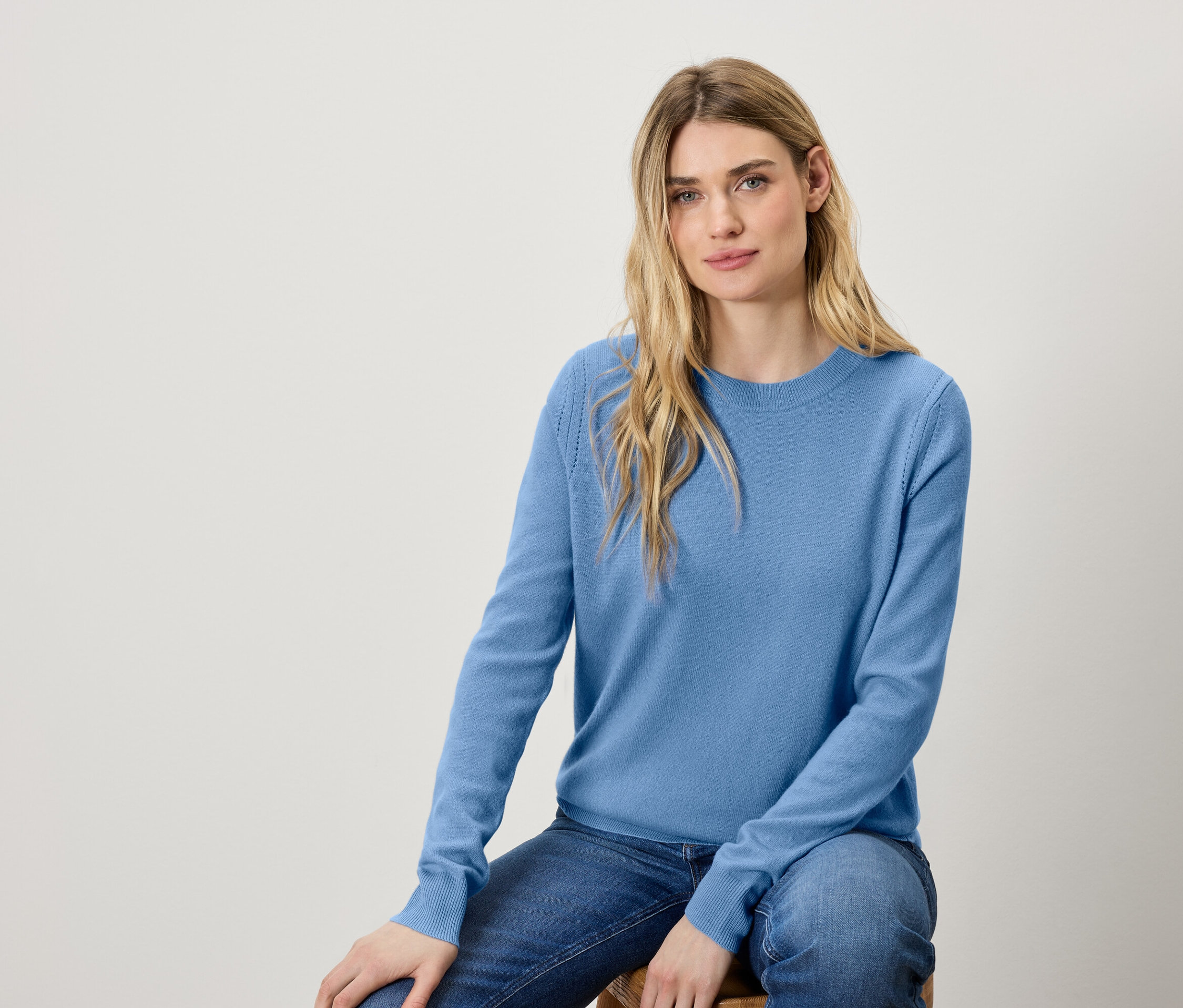 Une femme assise sur un tabouret, portant un pull bleu et un jean.