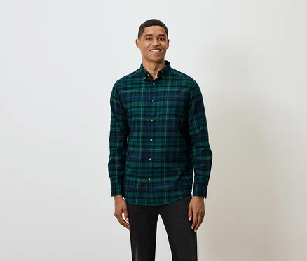 Un homme aux cheveux courts pose sur un fond blanc, portant une chemise à manches longues à carreaux verts et bleus et un jean noir.