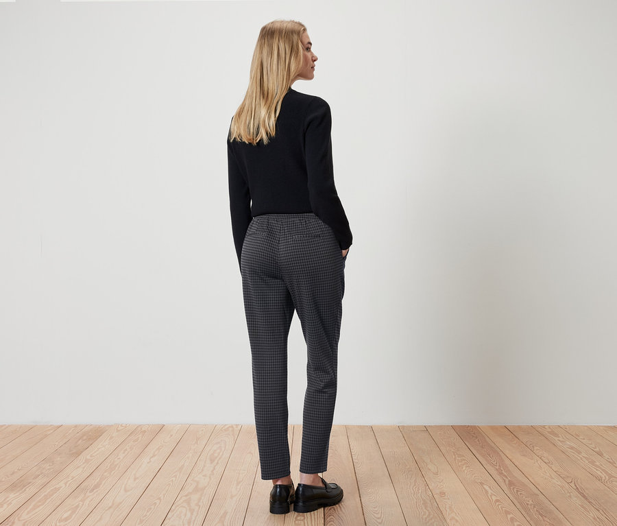 Une femme se tient dos à la caméra, portant un haut noir et un pantalon à carreaux gris.