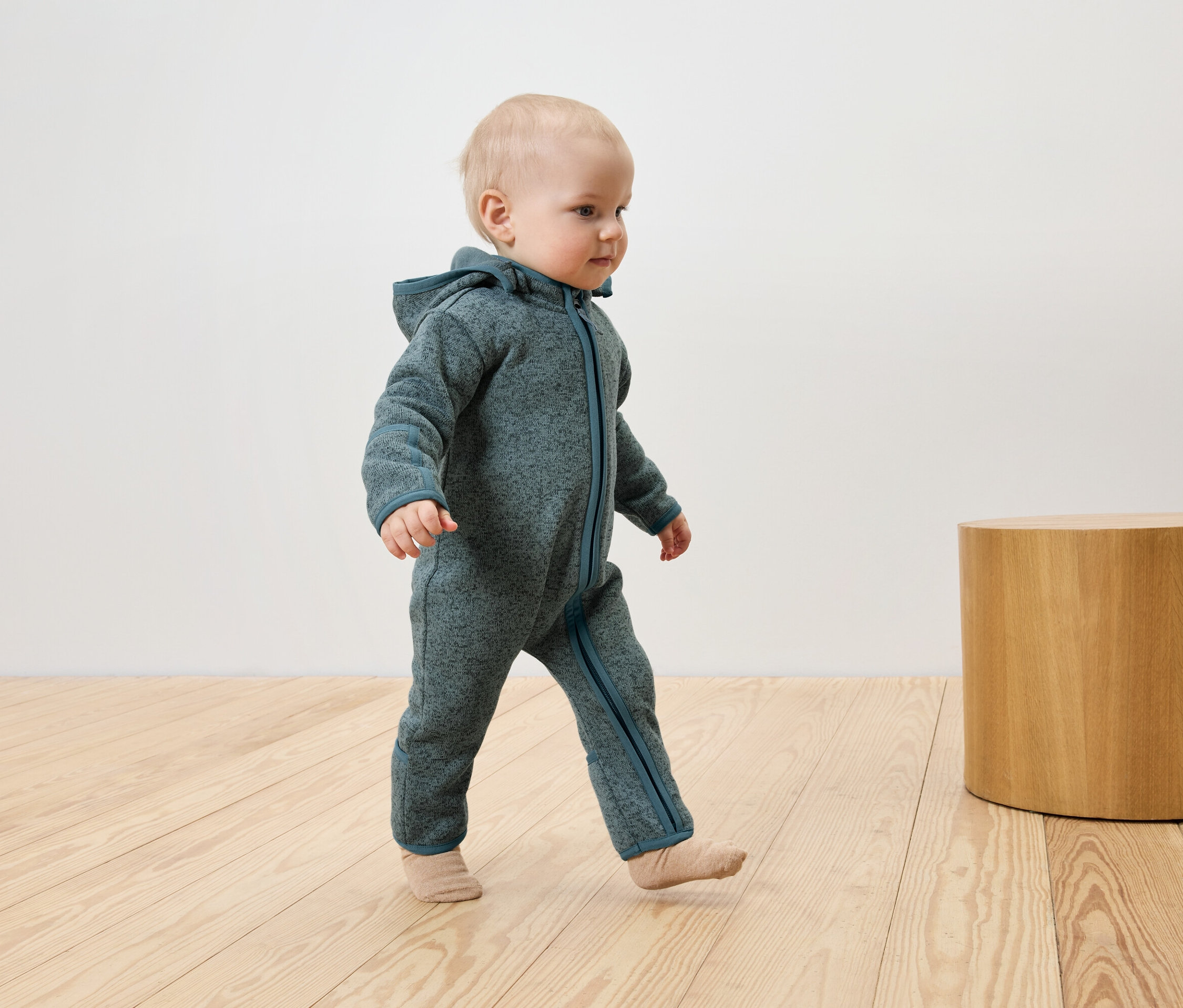 Un bébé vêtu d'une combinaison bleu-vert à capuche marche sur un plancher en bois à côté d'une petite table en bois.