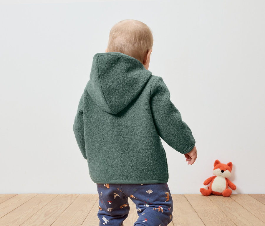 Un bébé, vu de dos, porte un manteau à capuche vert et un pantalon à motifs. Un renard en peluche est assis à côté de lui.