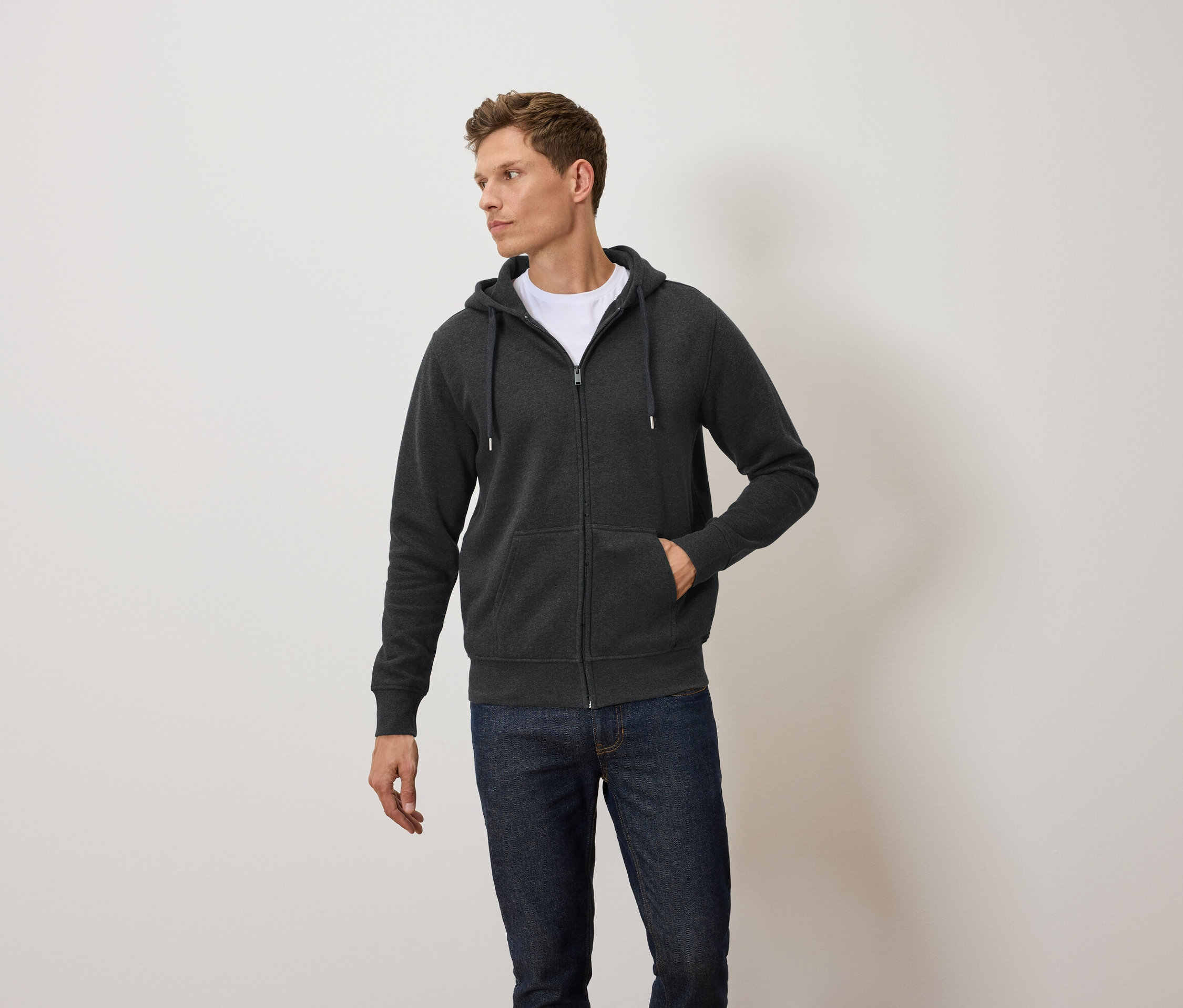 Un homme porte un sweat à capuche zippé gris foncé, un t-shirt blanc et un jean bleu.