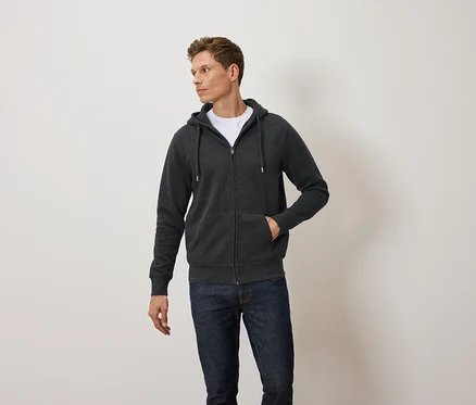 Un homme porte un sweat à capuche zippé gris foncé, un t-shirt blanc et un jean bleu.