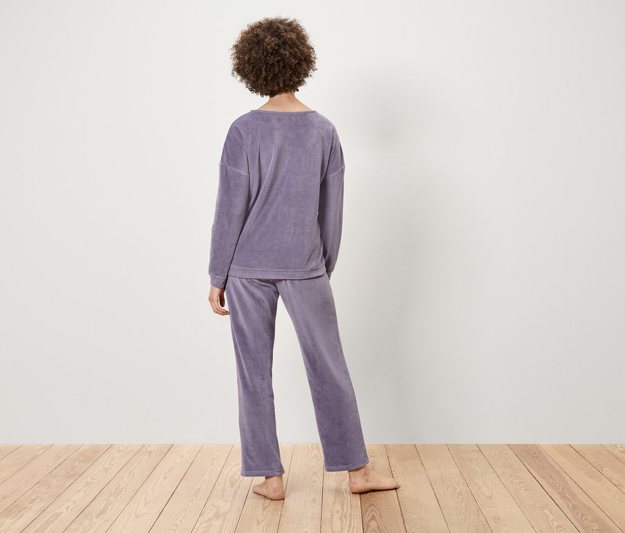 Une femme aux cheveux bouclés se tient dos à nous, portant un pyjama en velours violet sur une surface en bois clair.