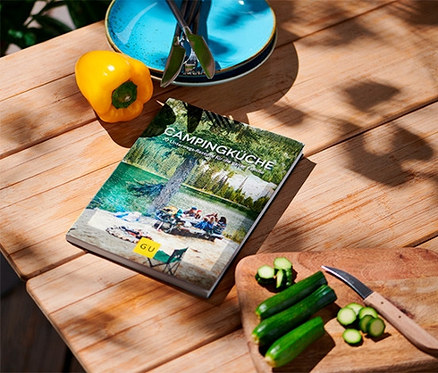 Sur une table en bois se trouvent le livre « Campingküche » (en allemand), un poivron jaune, des assiettes et des courgettes coupées.