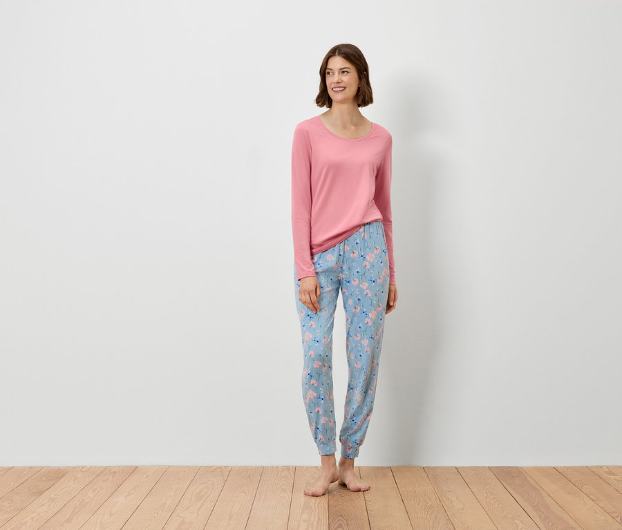 Une femme se tient debout et porte un haut rose à manches longues et un pantalon bleu à motifs floraux.