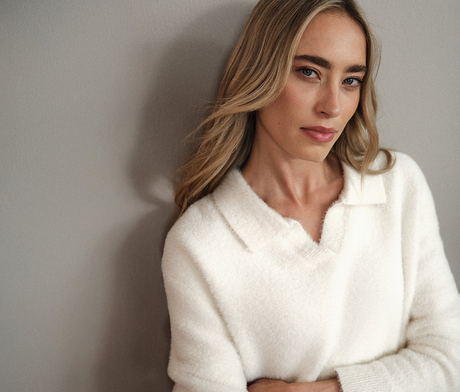 Une femme blonde portant un pull en maille à col polo Street One blanc est appuyée contre un mur gris.