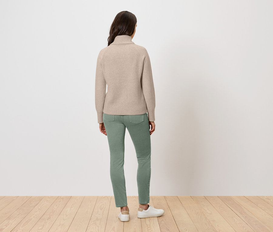 Une femme se tient dos à la caméra portant un pull à col roulé beige, un jean vert et des baskets blanches.