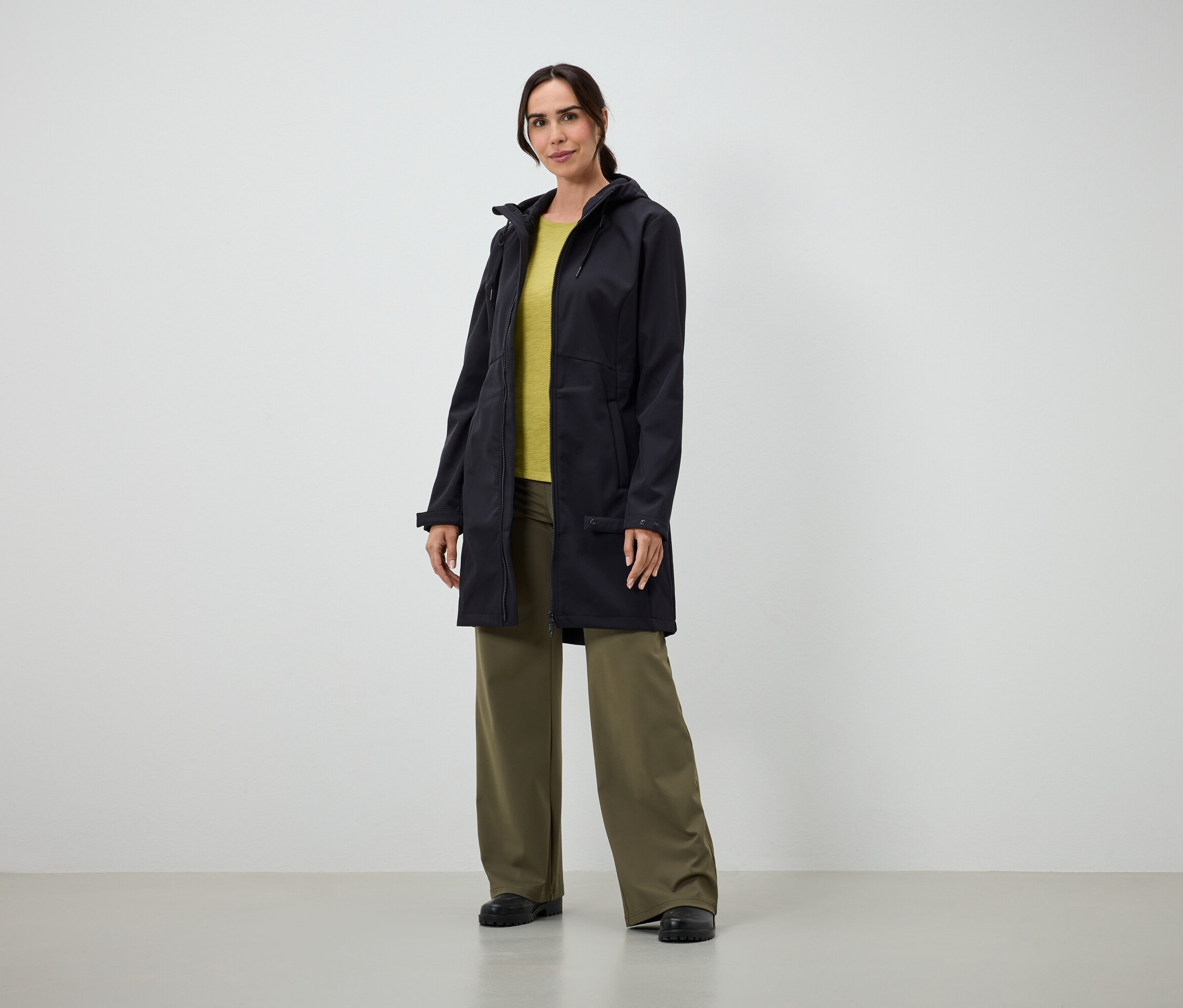 Une femme portant un long manteau noir, un t-shirt jaune et un pantalon vert ample se tient à l'intérieur.