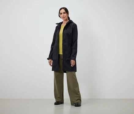 Une femme portant un long manteau noir, un t-shirt jaune et un pantalon vert ample se tient à l'intérieur.