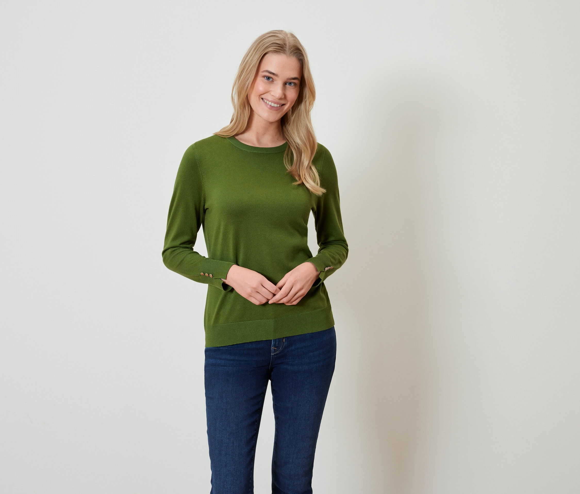 Une femme blonde se tient debout et porte un pull vert et un jean bleu.