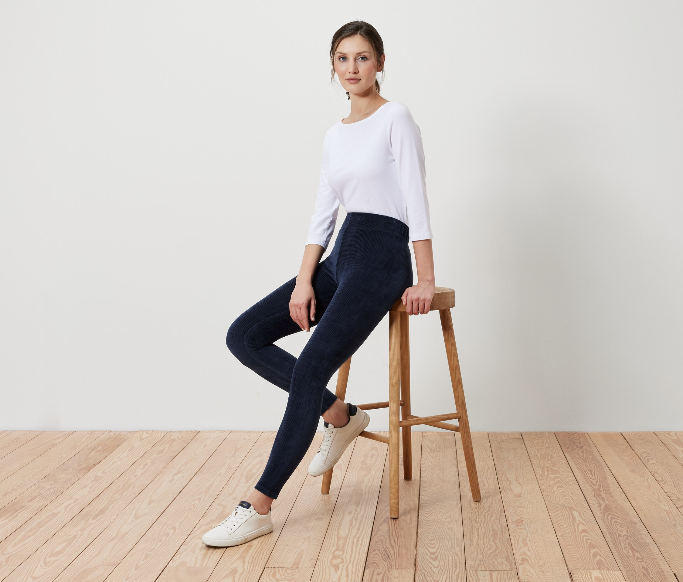 Une femme est assise sur un tabouret en bois, portant des leggings bleu marine et un t-shirt blanc.