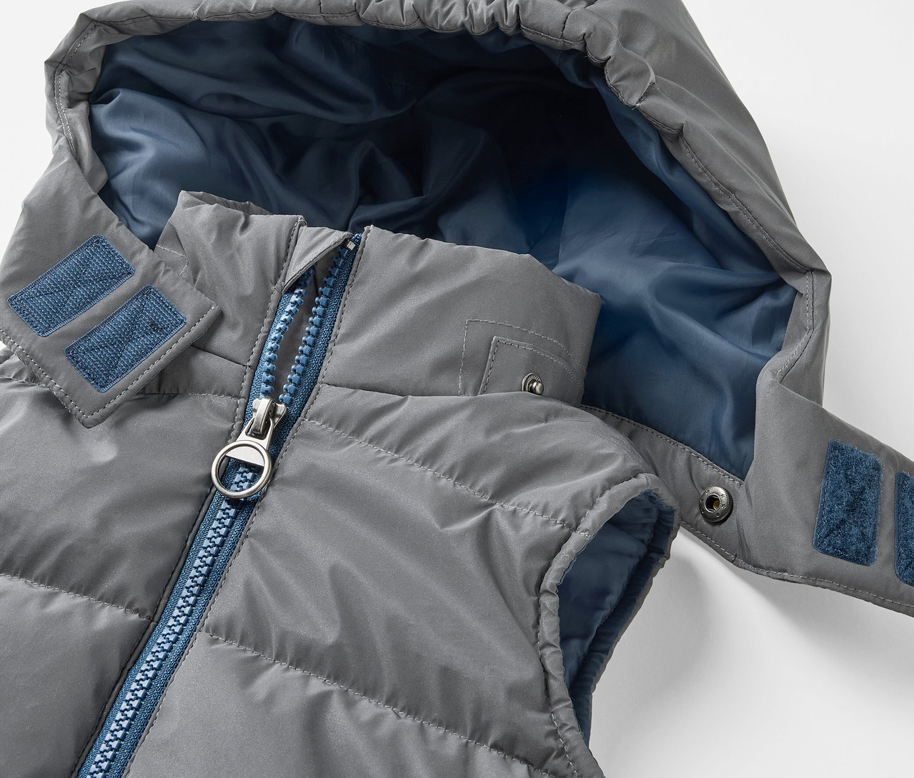 Gros plan sur une veste matelassée grise pour enfants avec capuche. La Veste matelassée réfléchissante pour enfants a une fermeture éclair bleue et un intérieur de capuche bleu.