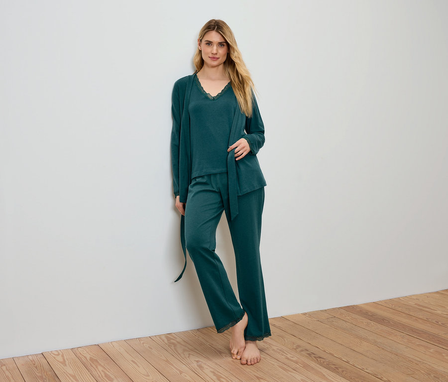 Une femme blonde en pyjama vert et peignoir appuyée contre un mur.