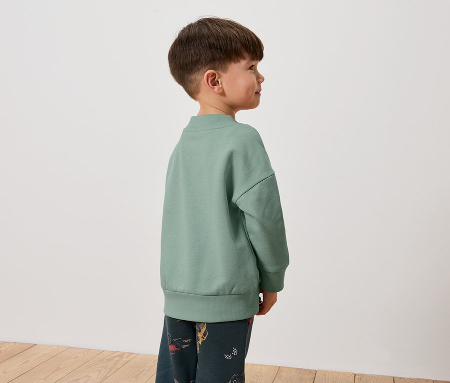 Un garçon portant un sweat-shirt vert et un pantalon à motifs se tient de profil.