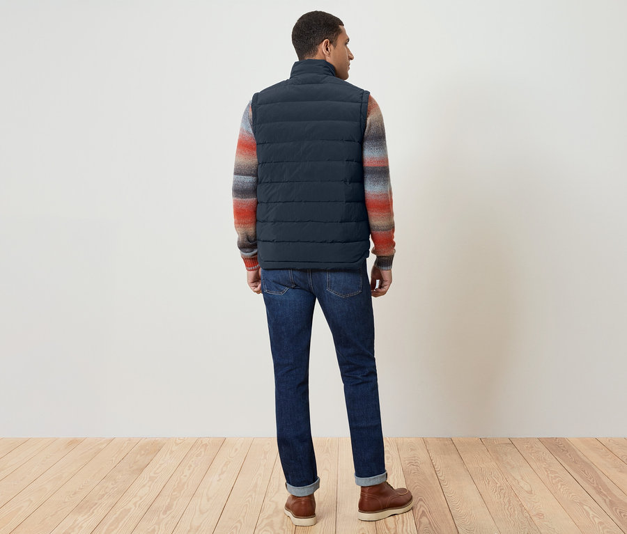 Un homme, dos à la caméra, porte un gilet bleu marine, un pull coloré, un jean et des chaussures marron.