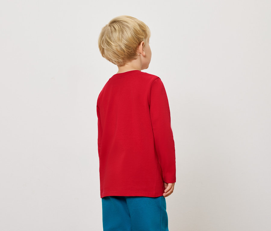 Un garçon blond, vu de dos, porte un t-shirt rouge à manches longues et un pantalon bleu.