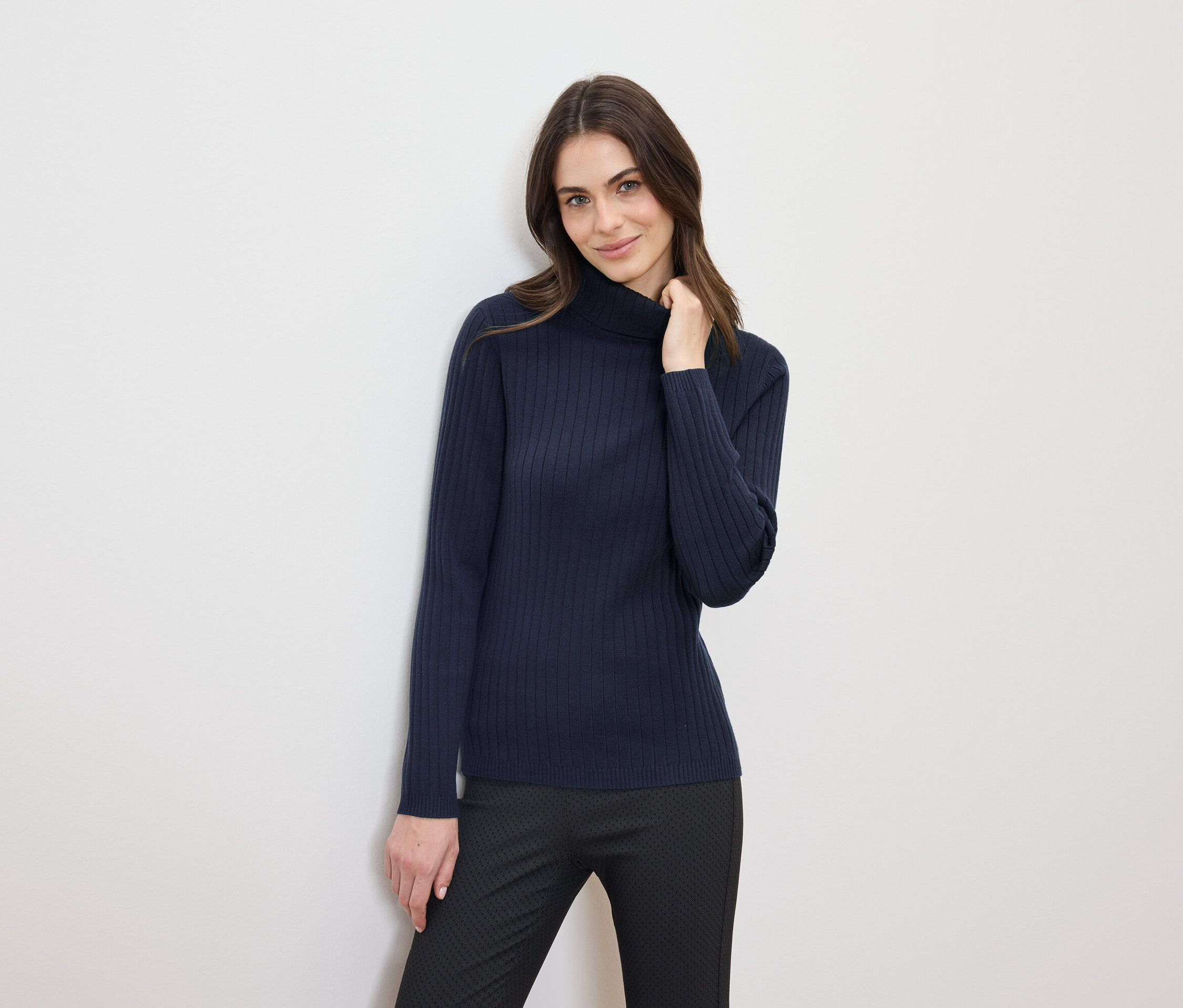 Une femme aux cheveux bruns pose dans un pull à col roulé bleu marine et un pantalon noir.