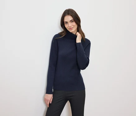 Une femme aux cheveux bruns pose dans un pull à col roulé bleu marine et un pantalon noir.