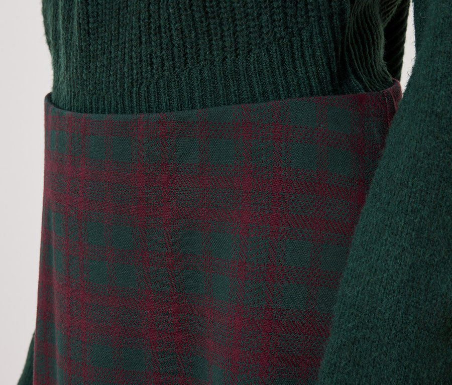 Détail d'une tenue féminine avec un pull vert et une jupe à carreaux.