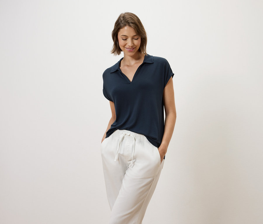 Femme aux cheveux bruns posant en polo bleu marine et pantalon blanc.
