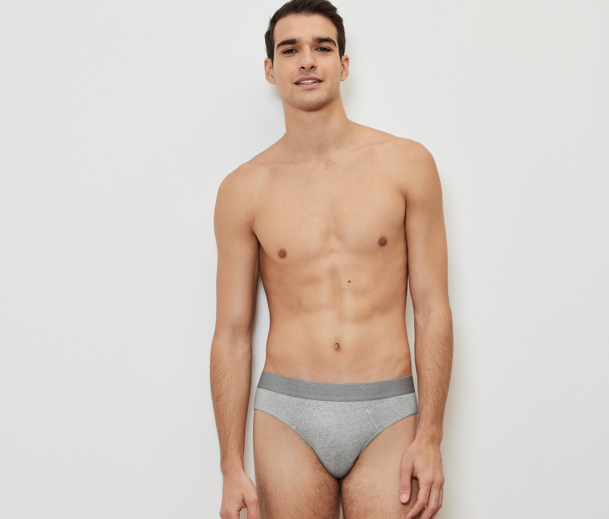 Un homme pose sur un fond blanc, portant un slip gris d'un lot de 4.