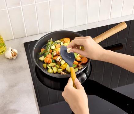 Une personne râpe une gousse d'ail dans une poêle de légumes sur une plaque de cuisson.
