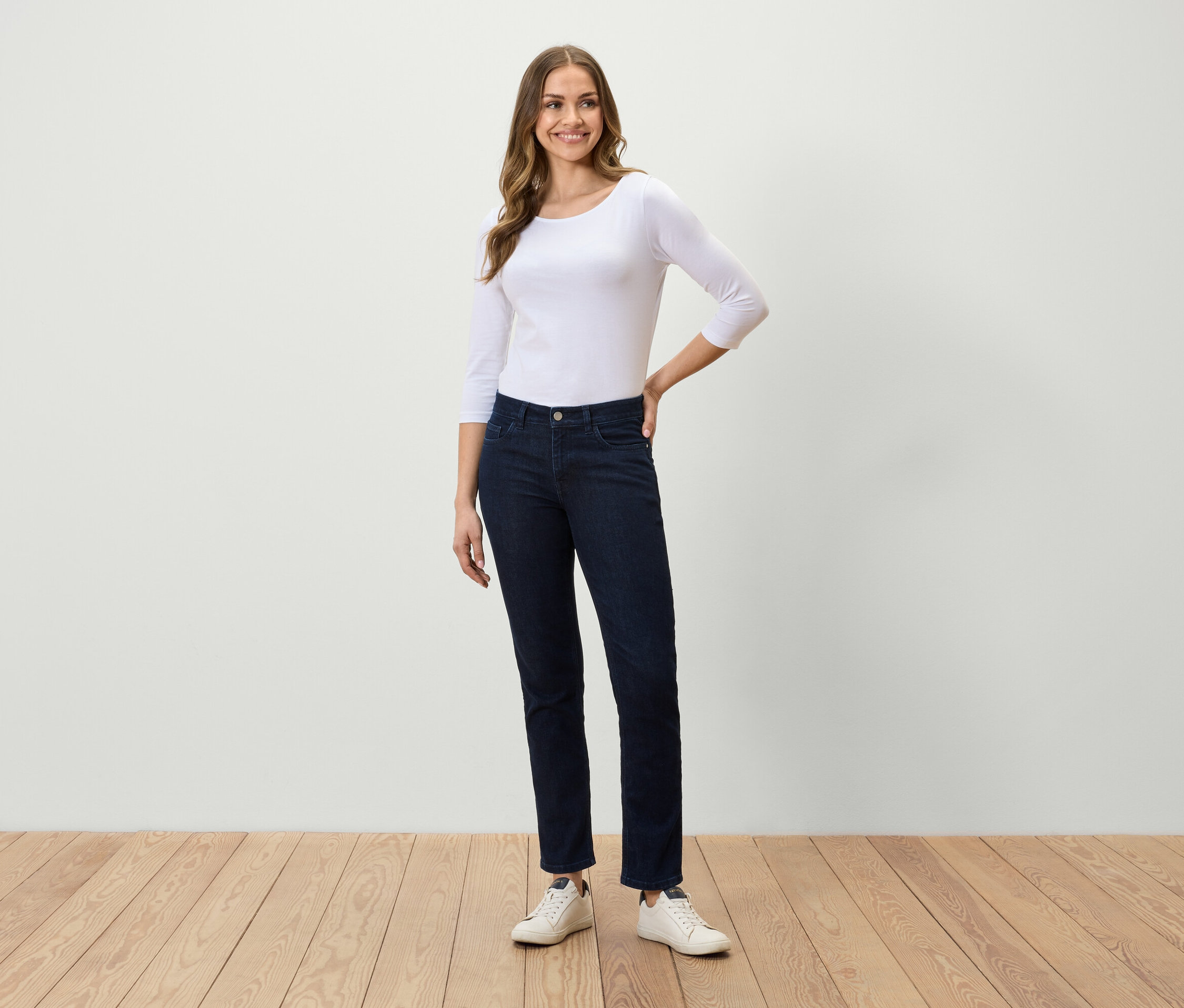 Une femme debout sur un plancher en bois porte un t-shirt blanc à manches trois quarts, un jean bleu foncé et des baskets blanches.