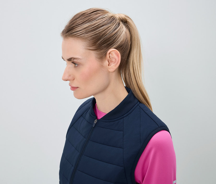 Profil d'une femme aux cheveux blonds attachés en queue de cheval, vêtue d'un gilet matelassé bleu marine et d'un haut rose.